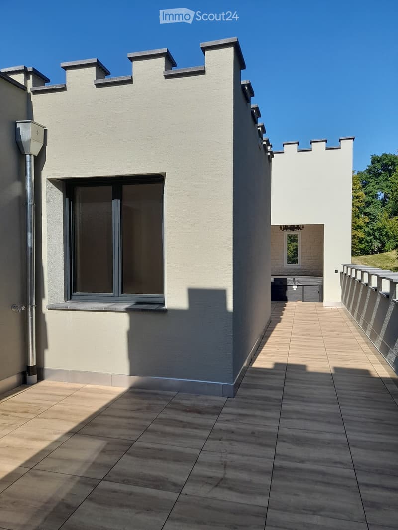 Modern balcony with wooden decking, grey stone walls, and a large window overlooking the area.