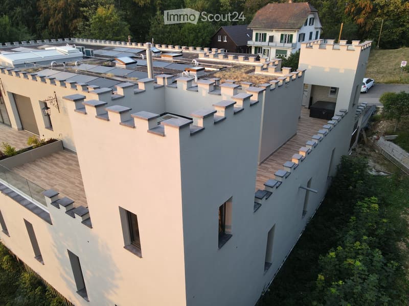 Modern two-story building with a distinctive, castle-like design featuring crenellated roofline and multiple chimneys. The structure includes a wooden deck area, glass railing, and surrounding greenery.