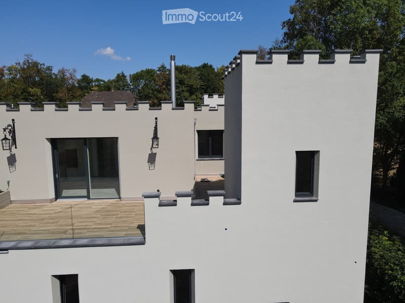 Modern two-story house with white exterior, crenellated roofline, large windows, and a balcony area with wooden decking.