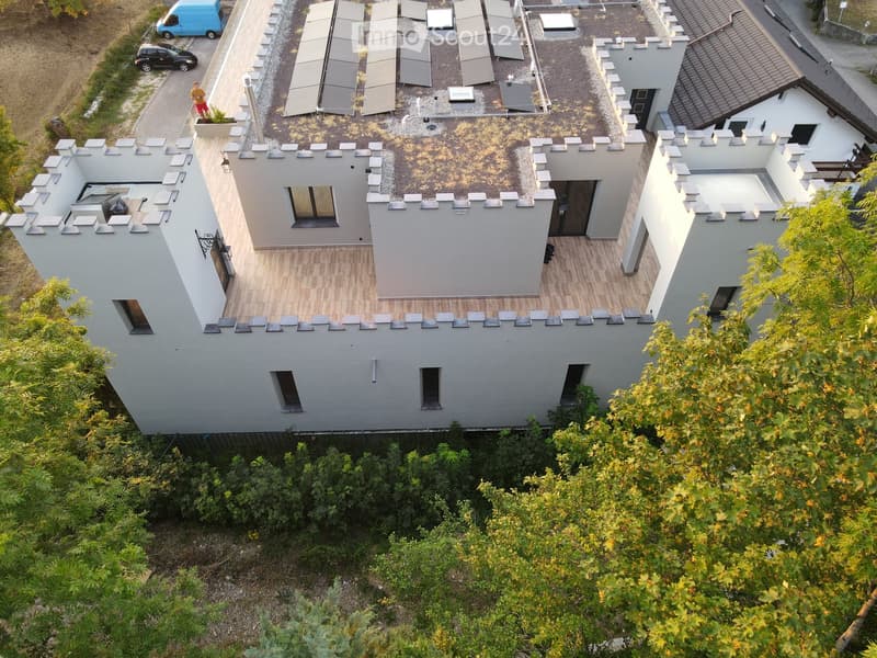 Modern two-story building with a flat roof and decorative battlements, surrounded by greenery and trees.