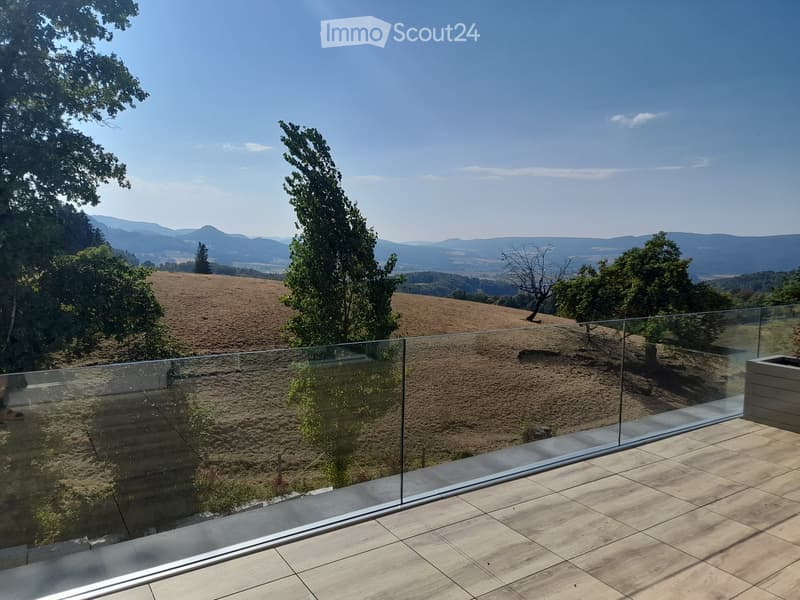 A modern glass-enclosed balcony with a panoramic view of rolling hills and a clear blue sky.