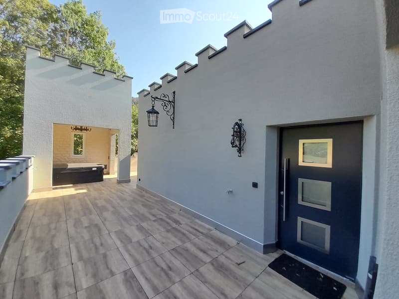 A modern outdoor area with tiled flooring, a grey front door, decorative wall-mounted lanterns, and a small seating area with a black bench and a small table.