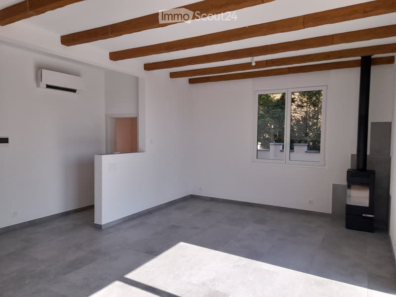 Empty room with white walls, exposed wooden beams, grey tiled floor, large window, and a black wood-burning stove.