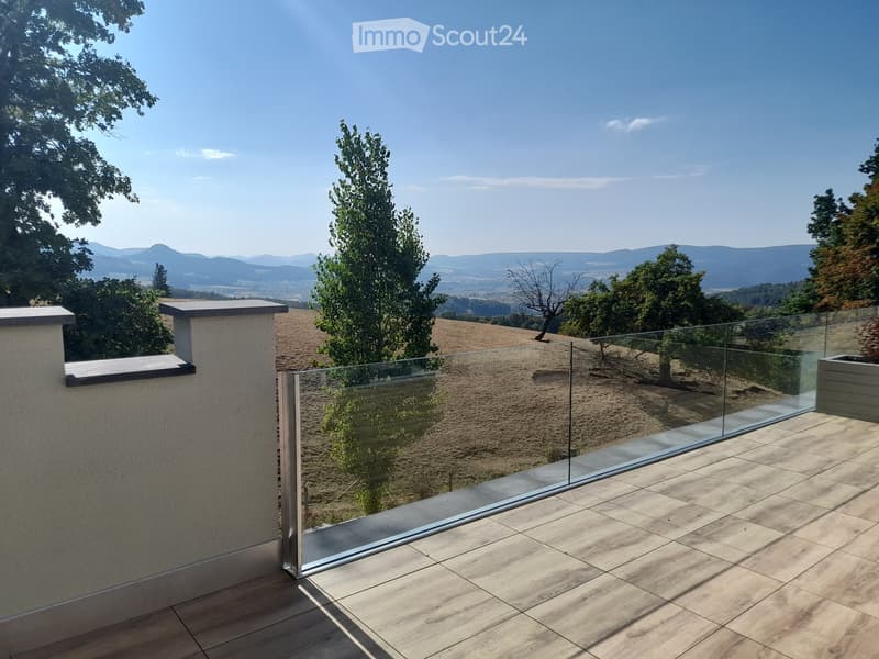 Modern glass-enclosed balcony with wooden decking, overlooking a scenic countryside with trees and distant hills.