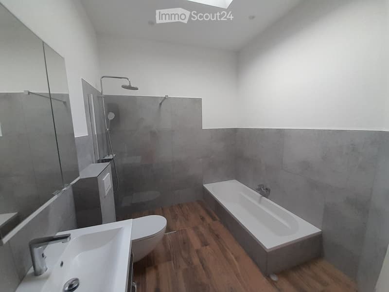 Modern bathroom with wooden flooring, grey tiled walls, a bathtub, toilet, sink, and a glass-enclosed shower area.