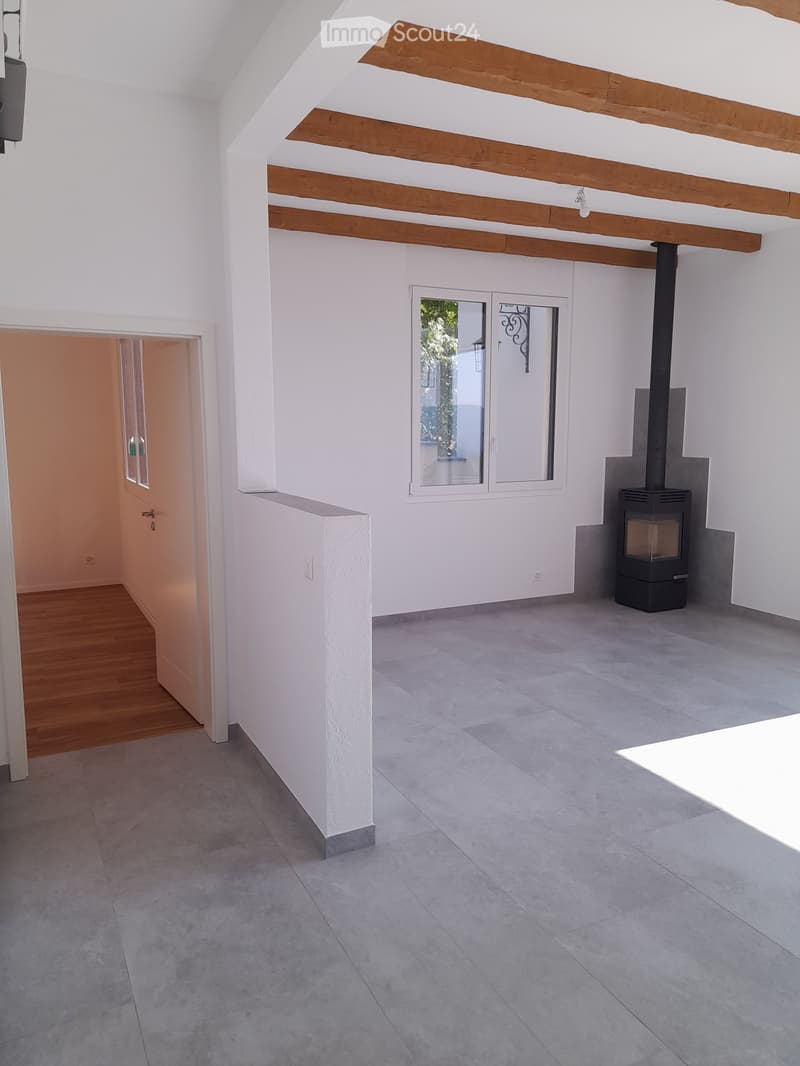 Empty room with exposed wooden beams, grey tiled floor, white walls, and a black wood-burning stove. There is a doorway leading to another room and a window with a view of greenery.