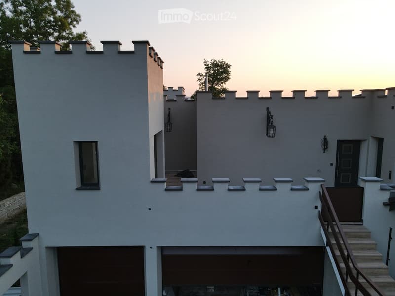 Modern two-story house with white walls, grey accents, and a raised ground floor. Features include a balcony, garage, and decorative wall lights.