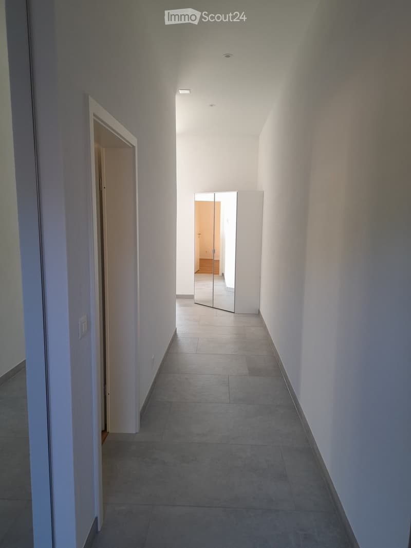 Empty hallway with tiled floor, white walls, and recessed lighting. Features a mirrored cabinet and an open doorway leading to another room.
