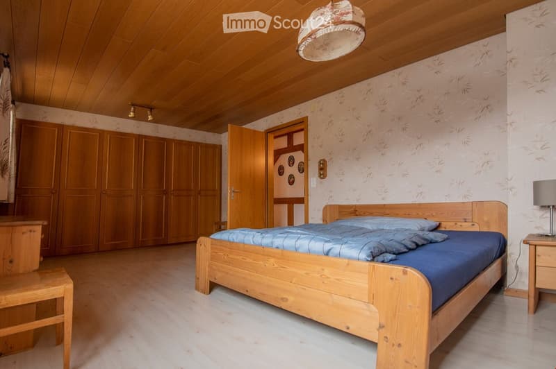Cozy bedroom with wooden bed, nightstand, wardrobe, and a small wooden stool. Light wooden ceiling and walls with floral wallpaper.