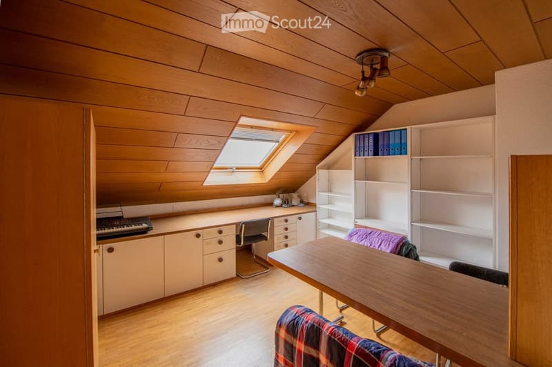 Bright attic room with wooden ceiling and floor, large skylight window, built-in desk with keyboard, shelves, and a small table with chairs.
