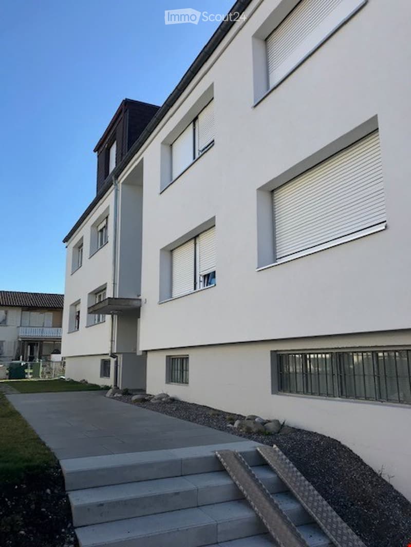 White painted building, with multiple windows, modern design, concrete pathway leading to the entrance, metal grills on the ground floor windows, a small roof over the entrance, gravel driveway, concrete steps leading to the entrance.