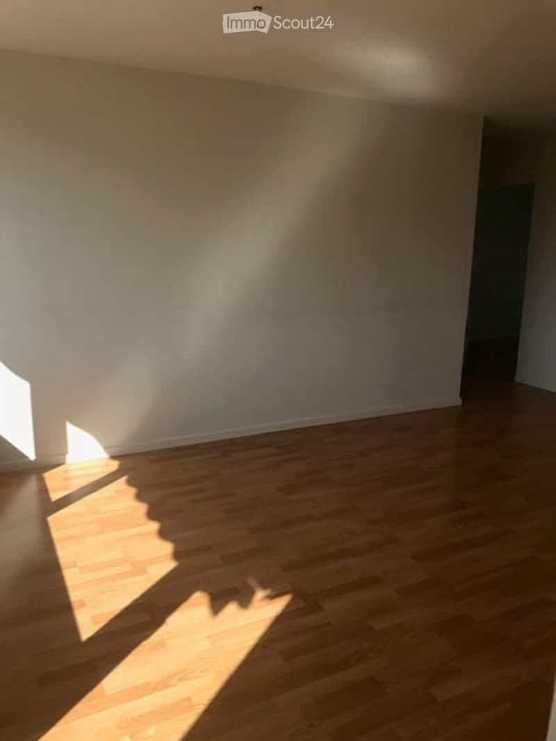 Empty room with wooden flooring and white walls, illuminated by natural light from a window on the left side.