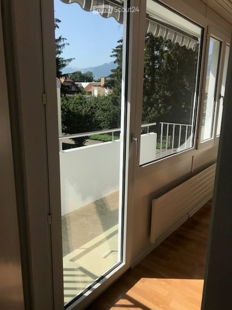 Balcony with white railing, overlooking a green area with trees and distant mountains.