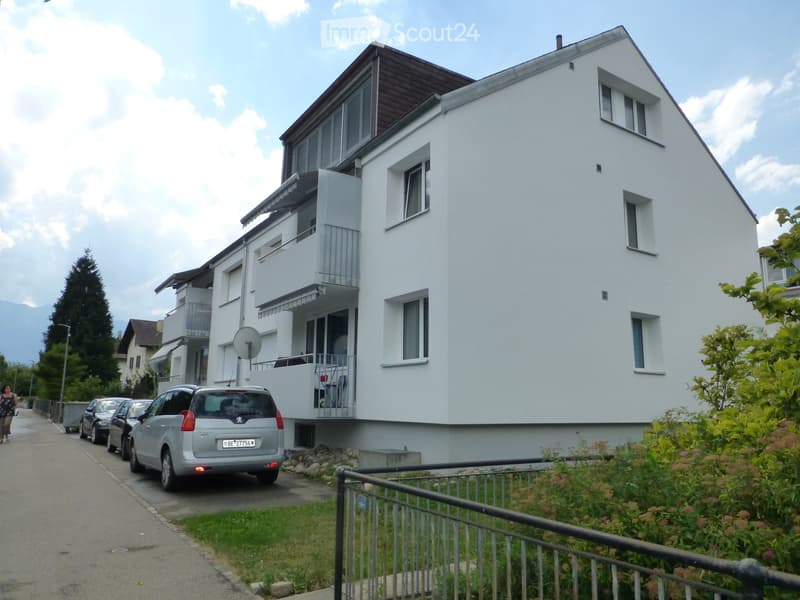 White exterior with balconies, three-story with two balconies, modern design, cars parked in front, surrounded by greenery.