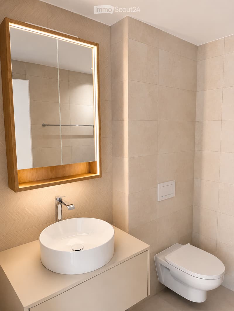 Modern bathroom with a white sink, sleek faucet, large mirror with wooden frame, and a toilet. Light beige tiles on walls and floor.