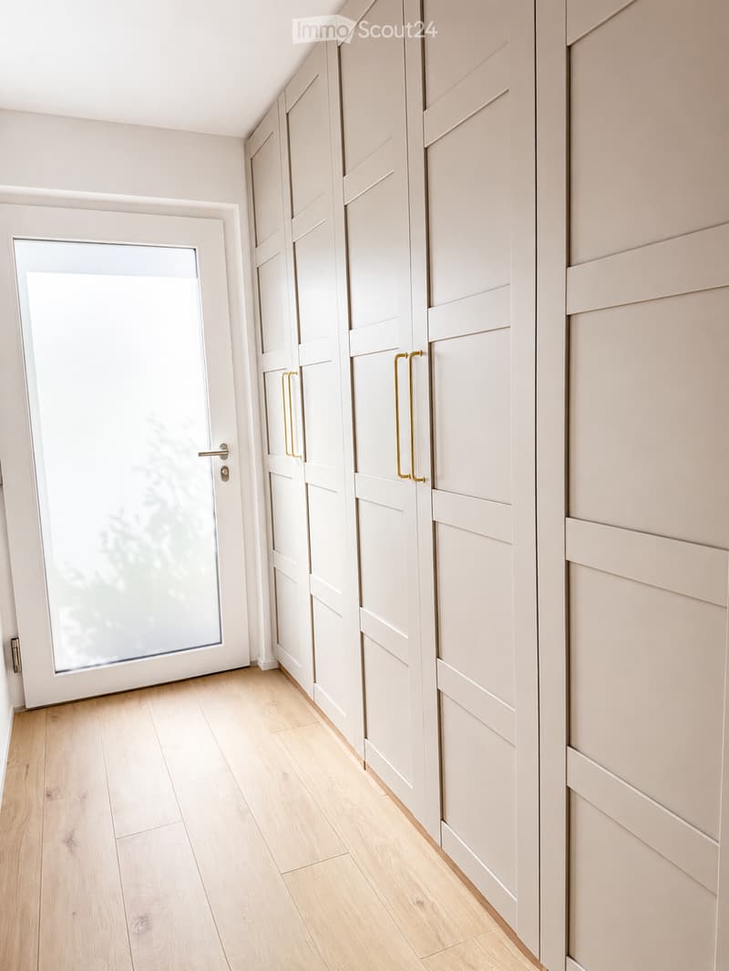 A clean, modern hallway with light wooden flooring, white walls, and built-in storage units with sleek, gold-colored handles. A frosted glass door is visible on the left side.