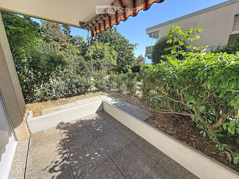 Balcony with tiled floor, small garden area with plants and shrubs, striped awning, and a view of surrounding greenery and buildings.