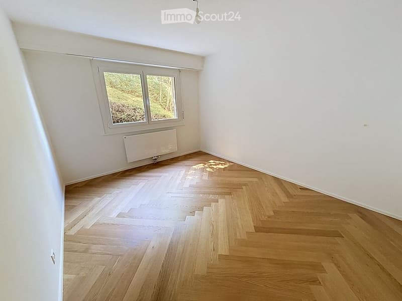 Empty room with wooden herringbone flooring, white walls, and a large window with a view of greenery. A radiator is mounted below the window.
