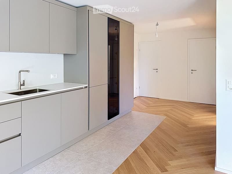 Empty room with light grey walls, wooden herringbone flooring, and a small kitchen area with white cabinets and a sink.