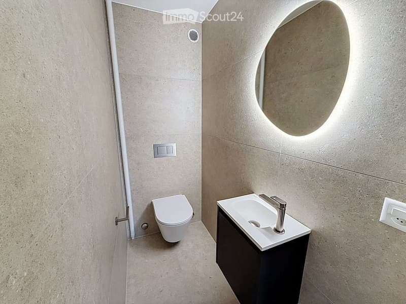 Modern bathroom with a toilet, sink, and a large, round mirror illuminated by ambient lighting. The walls and floor are covered in light beige tiles, and the sink features a sleek, contemporary design with a chrome faucet.