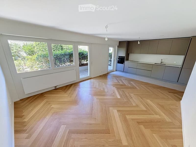 Bright, unfurnished room with wooden herringbone flooring, large windows, and a modern kitchen area with grey cabinets and stainless steel appliances.
