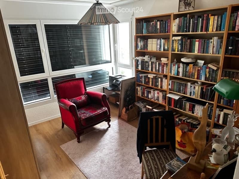 A cozy living room with large windows, a red armchair, a bookshelf filled with books, a small table, and a hanging lamp.