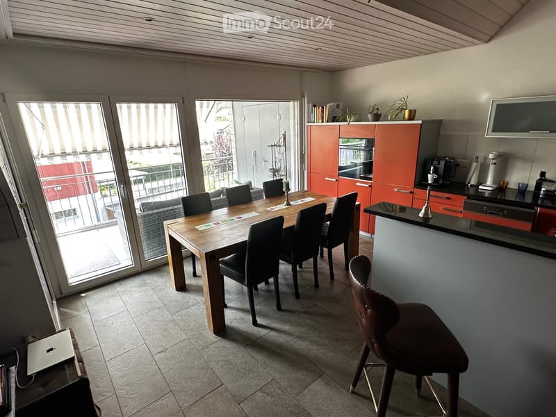 Open-plan living and dining area with a wooden dining table, black chairs, a grey sofa, and a modern kitchen with red cabinets and a coffee machine.
