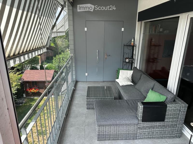 Balcony with grey wicker furniture, including a sofa, coffee table, and chair, with green cushions and a view of a garden.