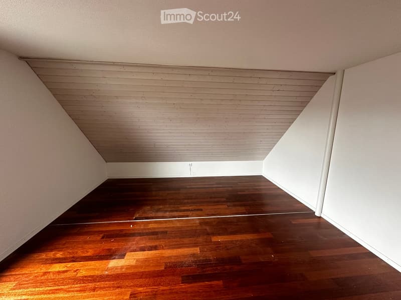 Empty room with wooden flooring and white walls, featuring a slanted ceiling with wooden paneling.
