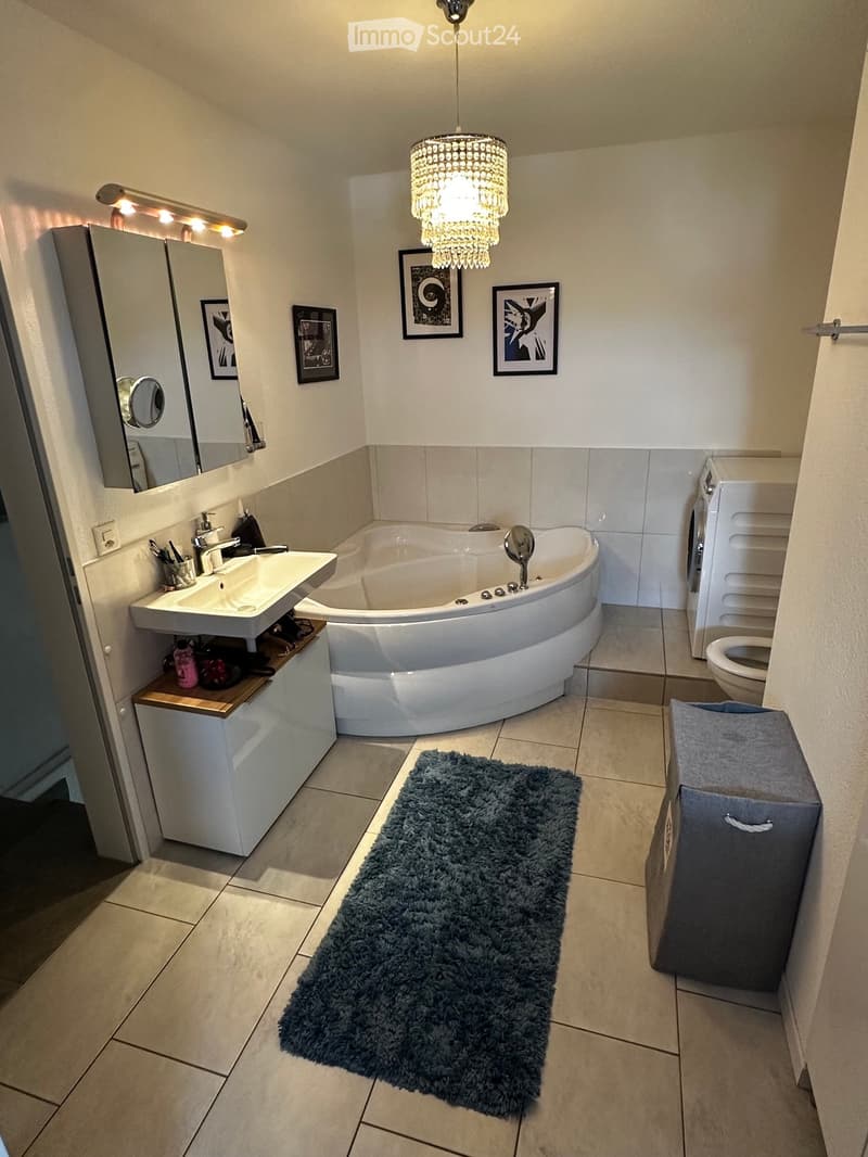 Modern bathroom with a freestanding bathtub, sink, toilet, and a large mirror. Features a fluffy blue rug, decorative picture frames, and a chandelier light fixture.