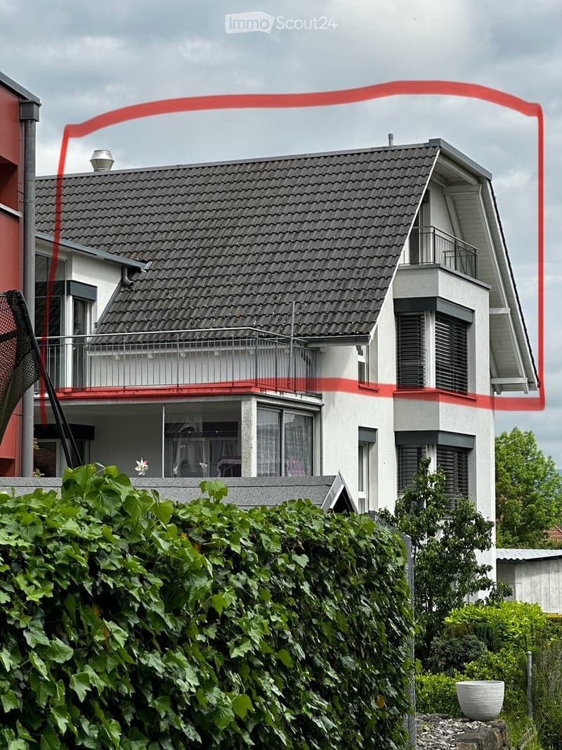 Two-story house with a gray tiled roof, white walls, and a balcony on the upper level. The house features multiple windows and is partially obscured by a dense green hedge in the foreground.