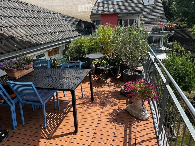 Balcony with wooden decking, metal table, blue chairs, potted plants, and a decorative sculpture.