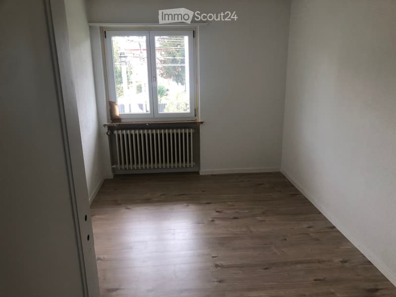 Empty room with wooden flooring, white walls, a window with white frame, and a radiator below the window.