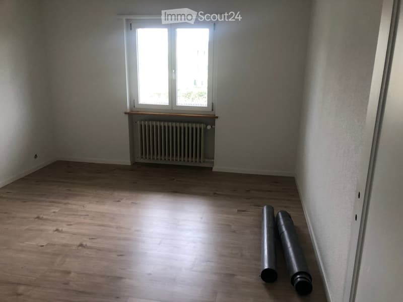 Empty room with wooden flooring, white walls, a window with a radiator below, and two large cylindrical objects on the floor.
