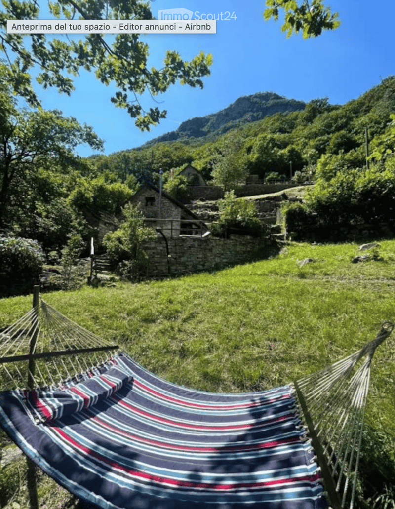 A serene outdoor setting with a hammock placed on a grassy area, surrounded by lush greenery and a stone house in the background, with a mountain view.