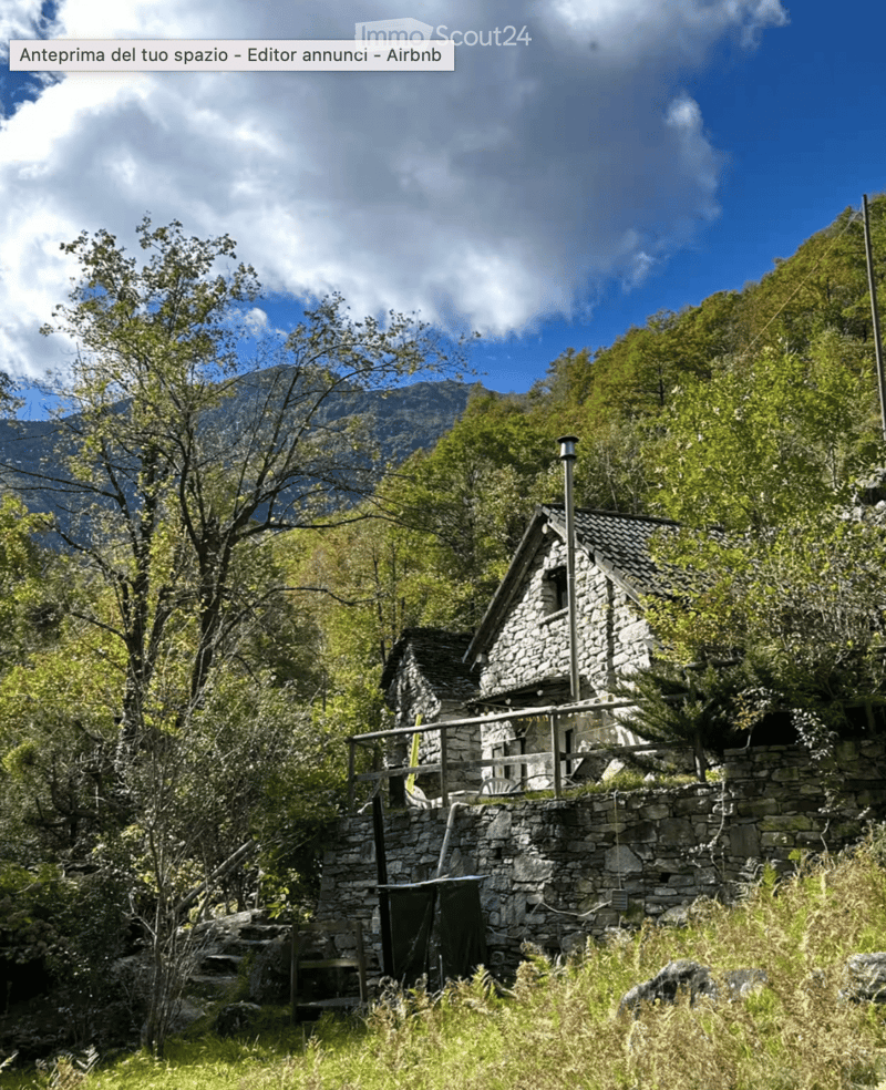A rustic stone house with a wooden balcony, surrounded by lush greenery and mountains in the background.