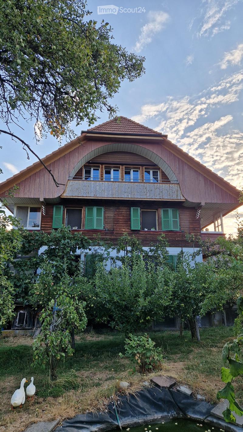 Two-story wooden house with green shutters, a balcony, and a surrounding garden with fruit trees and a small pond.