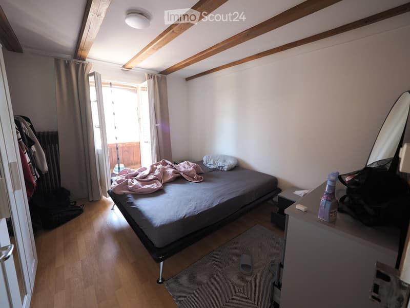 Cozy bedroom with wooden floor, exposed wooden beams, bed with grey duvet and pink blanket, nightstand with mirror, and a small rug on the floor.