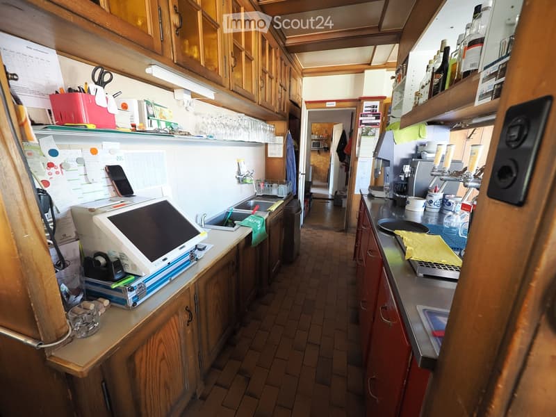The kitchen features wooden cabinets, stainless steel countertops, and various appliances including a laptop, microwave, and coffee machine. There is a sink with a faucet, and the space is organized with shelves holding glasses and other kitchen items.
