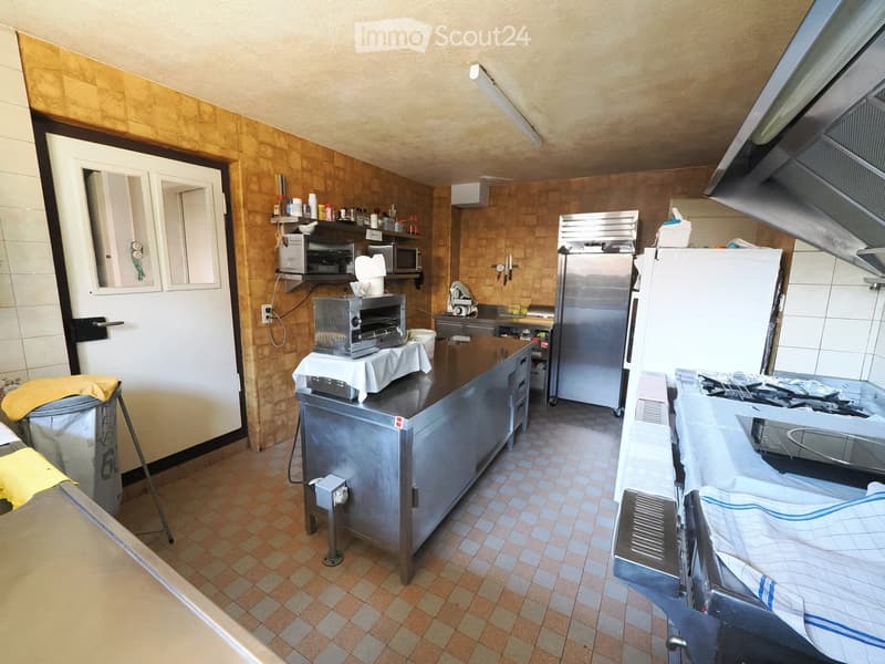 Commercial kitchen with stainless steel appliances, including a refrigerator, oven, microwave, toaster, and a large sink. Shelves hold various kitchen items, and a counter is visible.