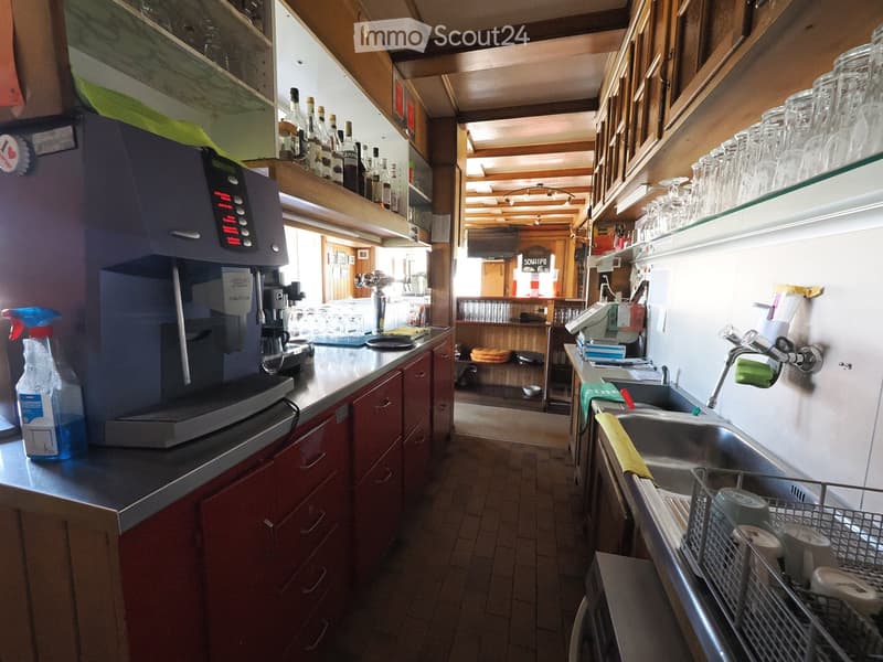 A small commercial kitchen with a coffee machine, sink, shelves with bottles and cups, and a dish rack.