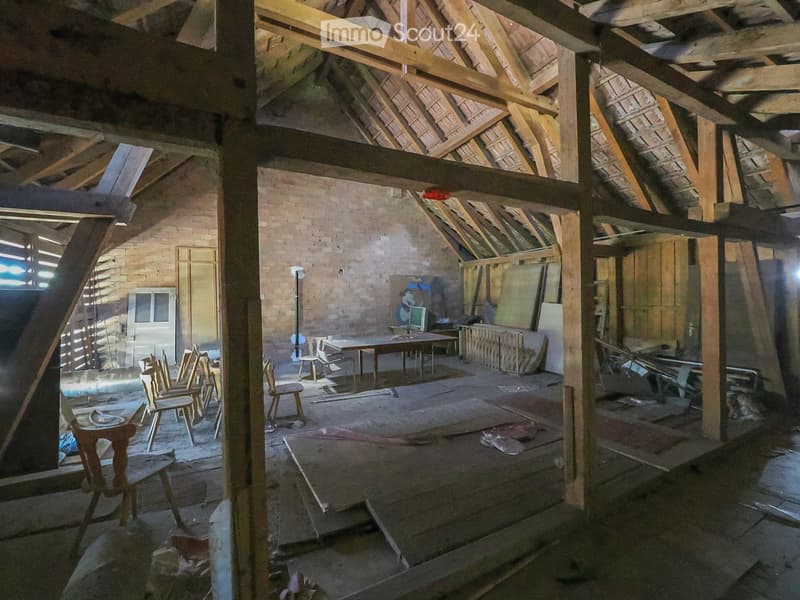 An attic space with exposed wooden beams and brick walls, featuring a few scattered chairs and a table, but largely empty and unfinished.