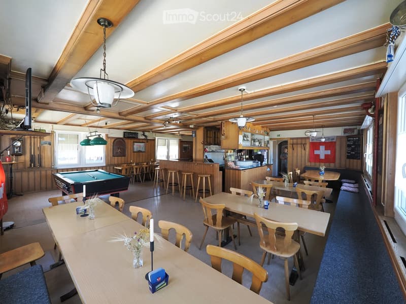Restaurant interior with wooden tables, chairs, stools, a pool table, and a bar area with shelves and stools.
