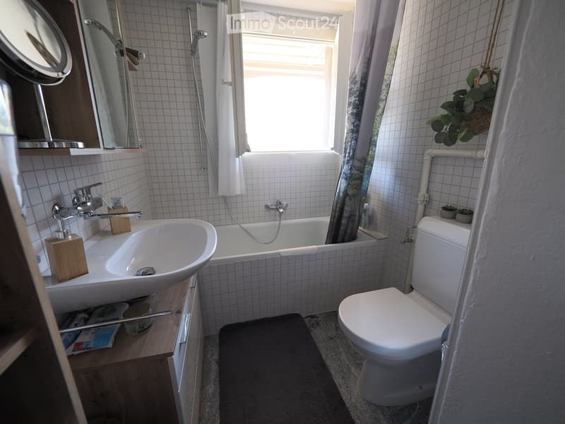 Bathroom with white tiled walls, a sink, bathtub, toilet, mirror, and a window with a curtain. Wooden shelf holds toiletries and a plant is on the windowsill.