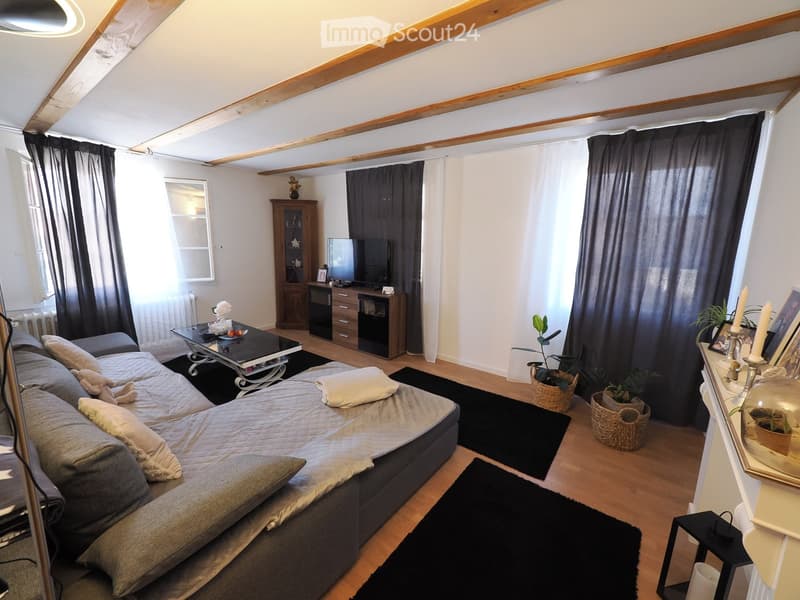 A cozy living room with a grey sectional sofa, coffee table, TV cabinet, and decorative plants. The room features wooden flooring, exposed wooden beams on the ceiling, and large windows with dark curtains.