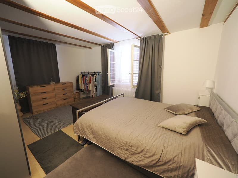 Cozy attic bedroom with wooden beams, a large bed, nightstand, dresser, and clothing rack. The room features exposed wooden beams, large windows with curtains, and a small table beside the bed.