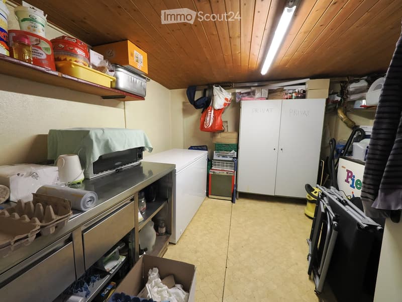 A cluttered utility room with a refrigerator, washing machine, shelves filled with various items, a vacuum cleaner, and a table with a covered appliance.