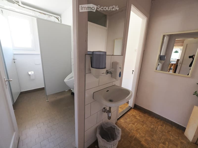 Clean bathroom with a sink, faucet, mirror, toilet, hand sanitizer dispenser, and a trash bin. The floor is tiled, and the walls are white.