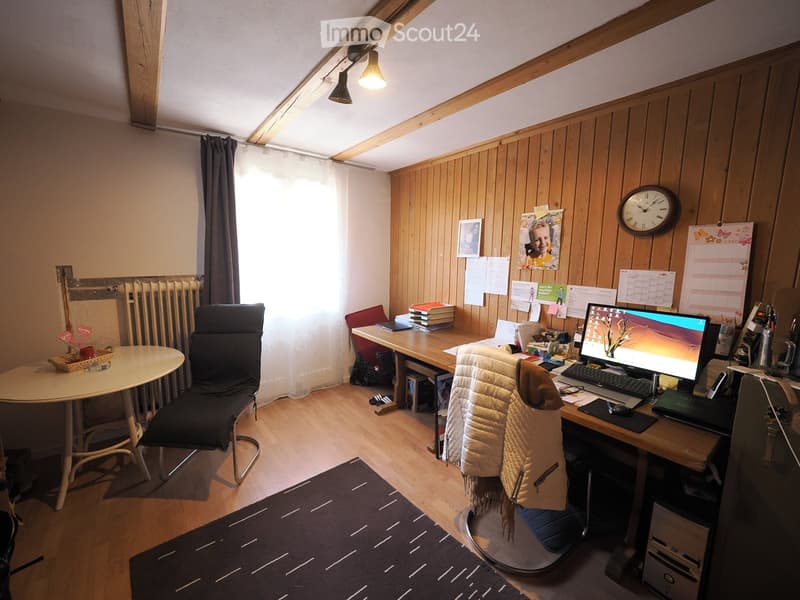 A cozy, well-lit room with wooden walls and a slanted ceiling. It features a desk with a computer setup, a round table with a chair, and a comfortable armchair with a blanket draped over it. The room includes a clock on the wall, picture frames, and a cal