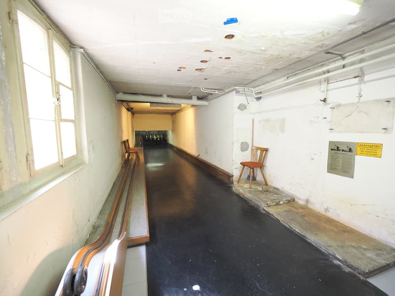 A long, narrow corridor with a dark floor, white walls, and a few chairs placed along the sides. The ceiling has exposed pipes and lighting fixtures.
